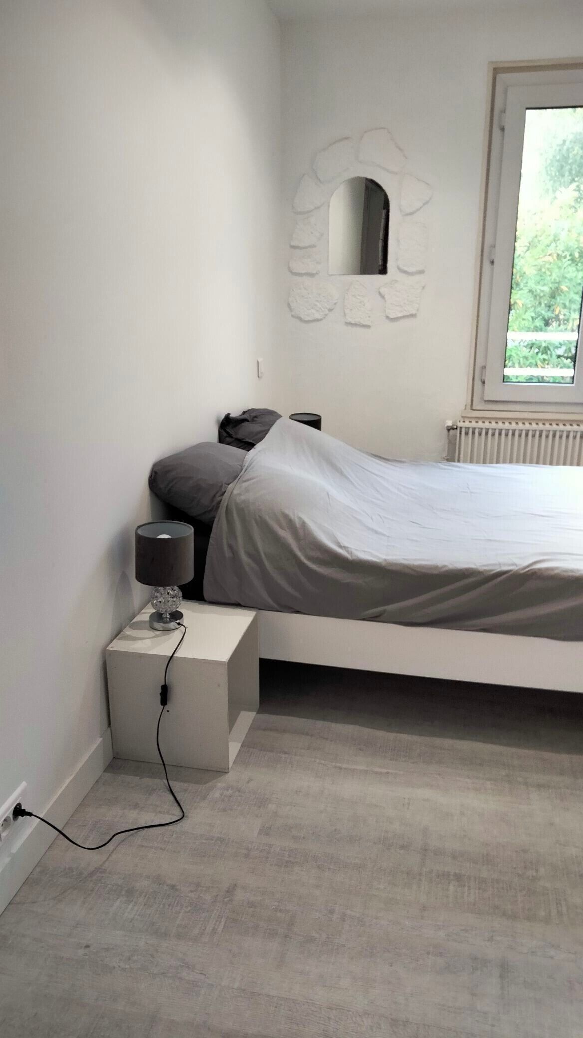 Une chambre à coucher avec un lit, une table de chevet avec une lampe, un miroir et une fenêtre.