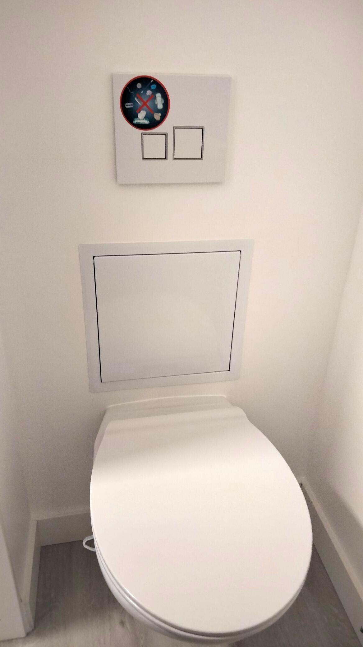 A white toilet with a flush handle, a wall-mounted flush button, and a small square vent.
