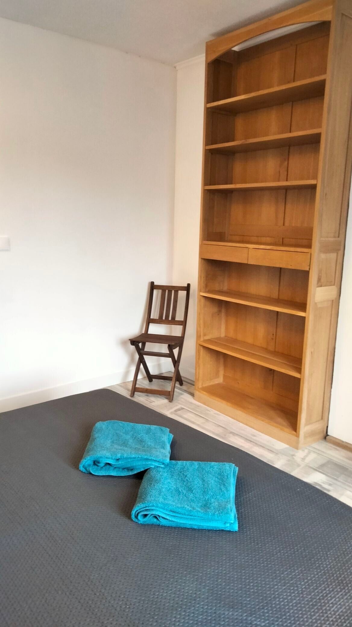 A room with a large wooden wardrobe, a small wooden chair, and two folded blue towels on a grey mat.