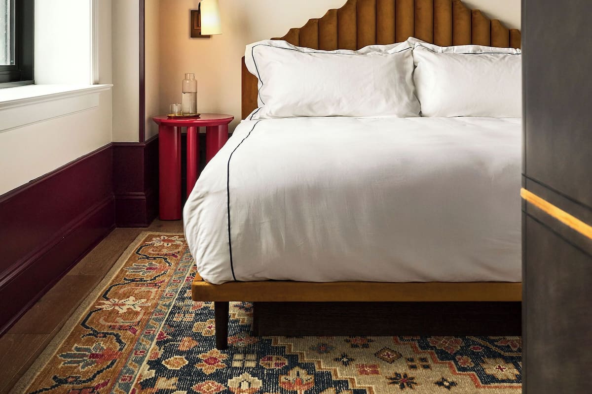 Una cama bien tendida con sábanas blancas, cabecera de madera y alfombra estampada.