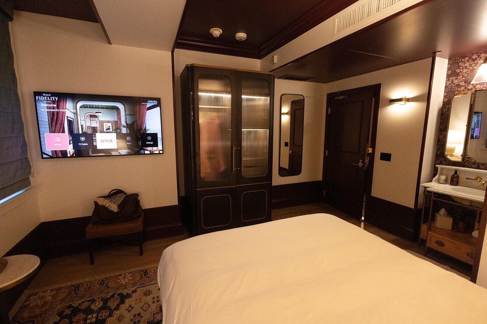 A hotel room with a large bed, a TV mounted on the wall, a wardrobe, a mirror, and a bathroom with a sink and mirror.