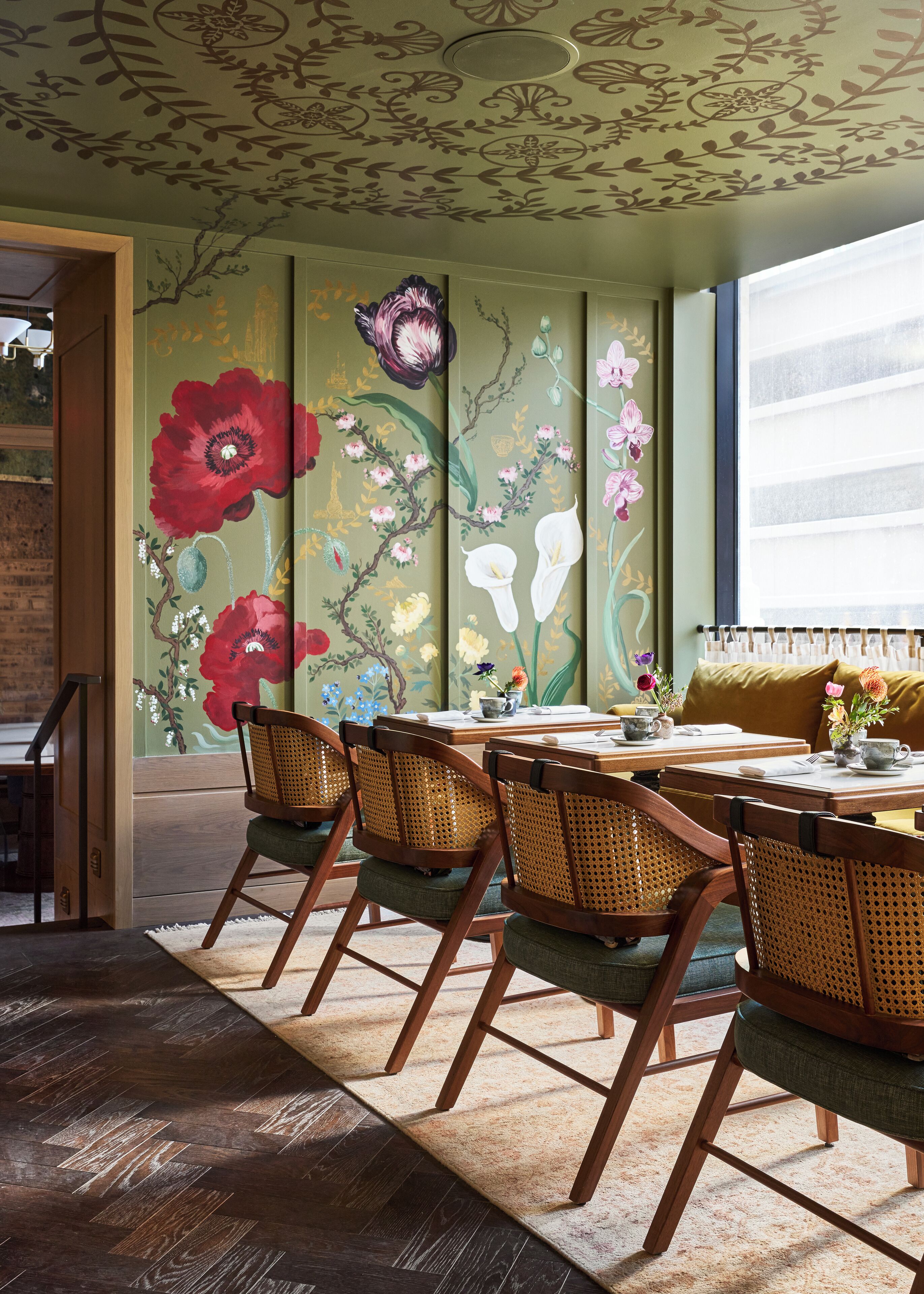 En matplats med ett bord dukat för fyra personer, omgiven av flätade stolar med gröna kuddar. Väggarna är dekorerade med blomstermotiv.