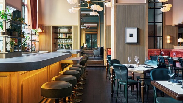 Un interno ristorante moderno con zona bar, soffitto alto con lampade a sospensione e area salotto centrale con tavoli e sedie.