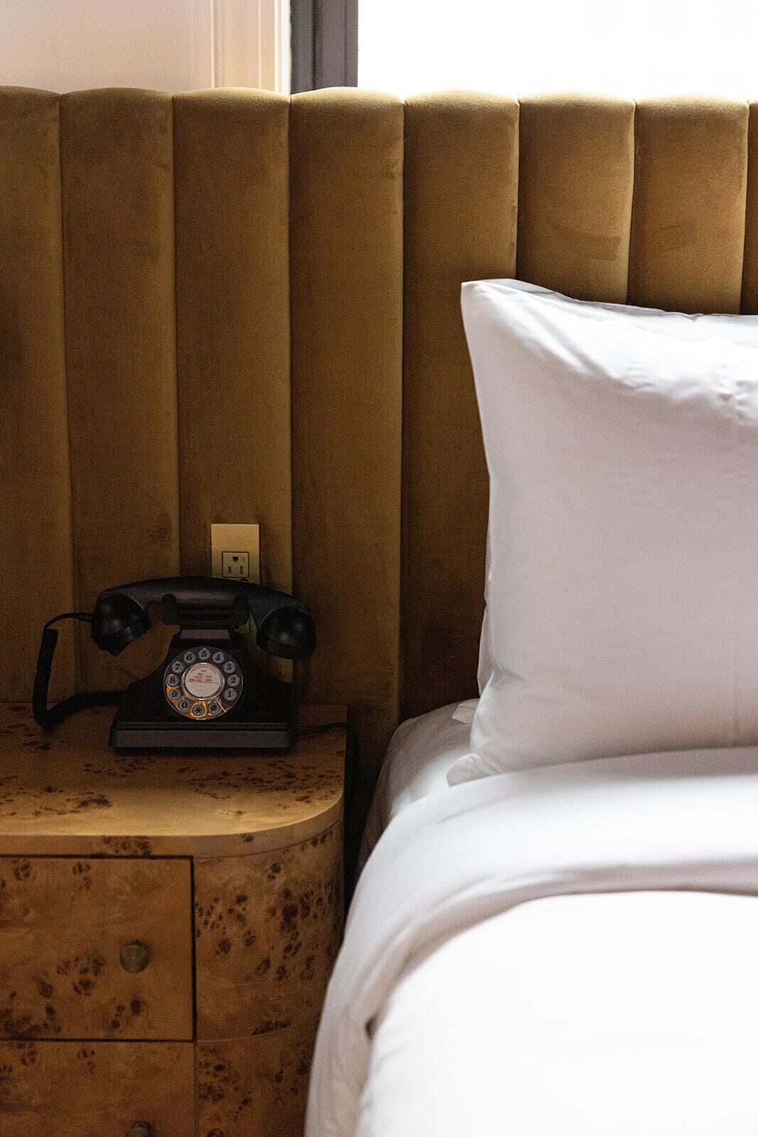 Een bed met een wit kussen en een oude draaischijf telefoon op een houten nachtkastje.