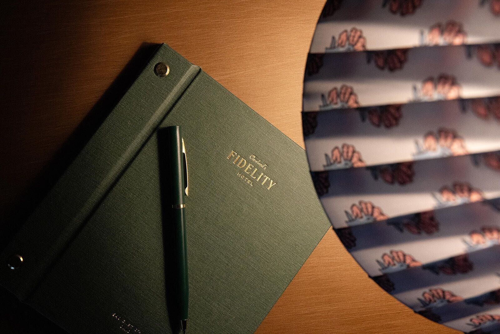 A green notebook with 'FIDELITY HOTEL' embossed on it, a pen, and a patterned paper with a floral design.
