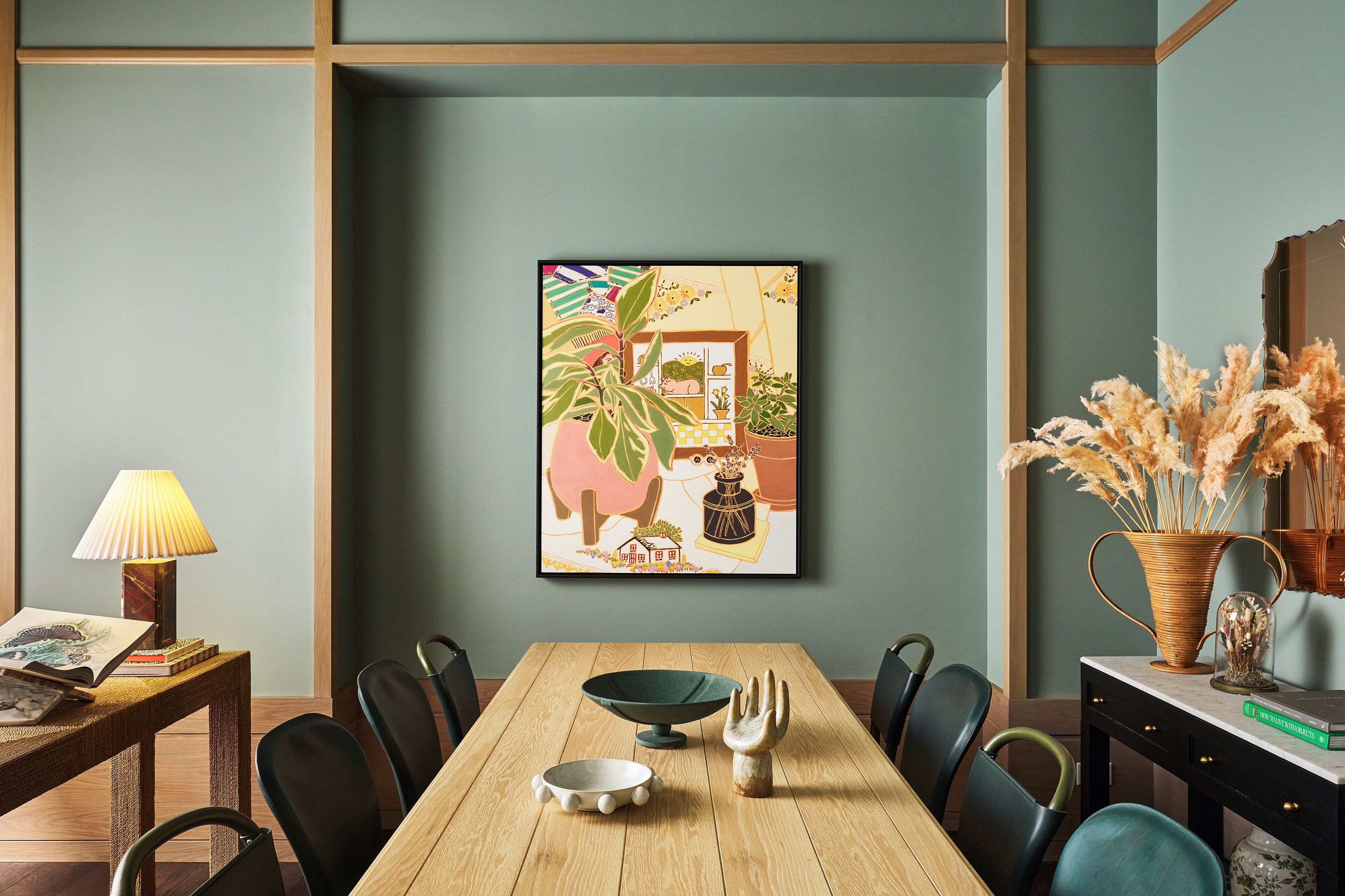 Uma área de refeições com uma mesa de madeira, cadeiras, uma obra de arte emoldurada, um abajur e um vaso com plantas secas.