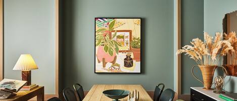 A dining area with a wooden table, chairs, a framed artwork, a lamp, and a vase with dried plants.