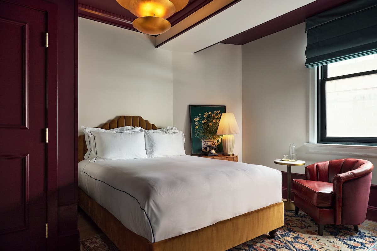 Habitación de hotel con una cama grande, una mesita de noche con lámpara, una silla y una ventana con vistas.
