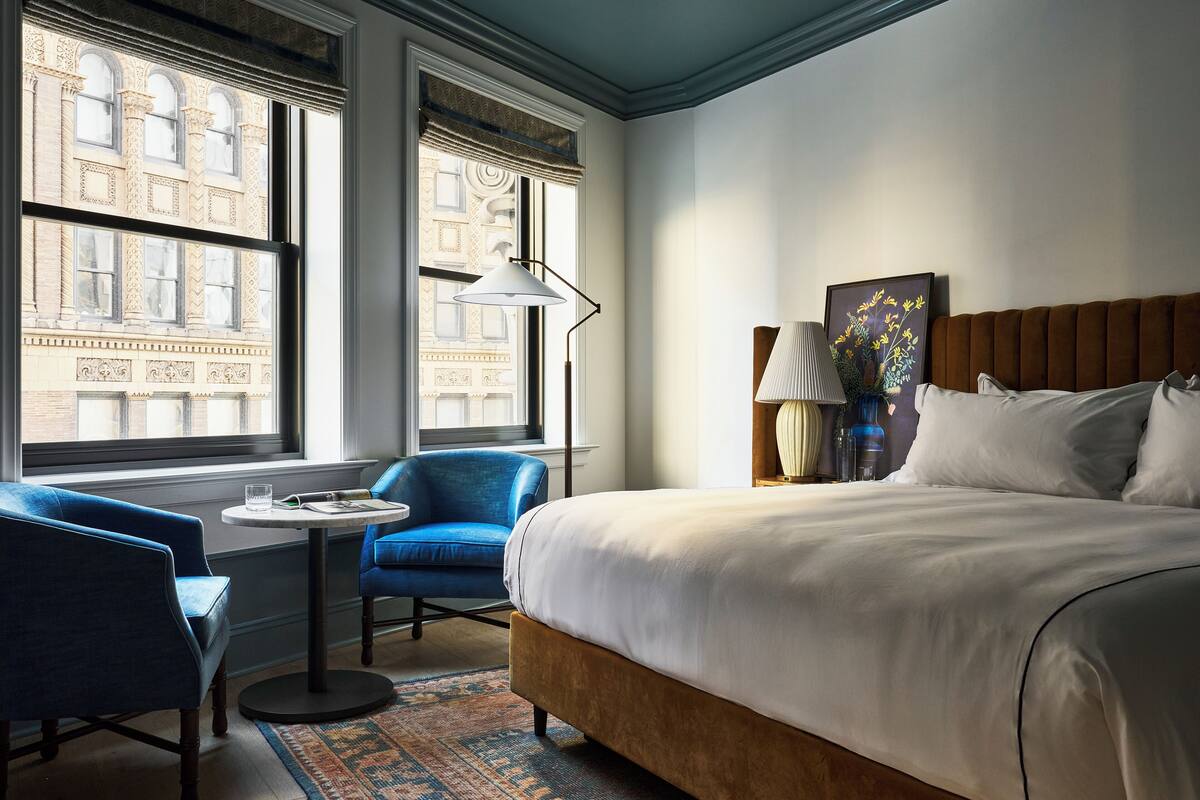 Una habitación de hotel con una cama grande, un sillón azul, una mesita y una lámpara.