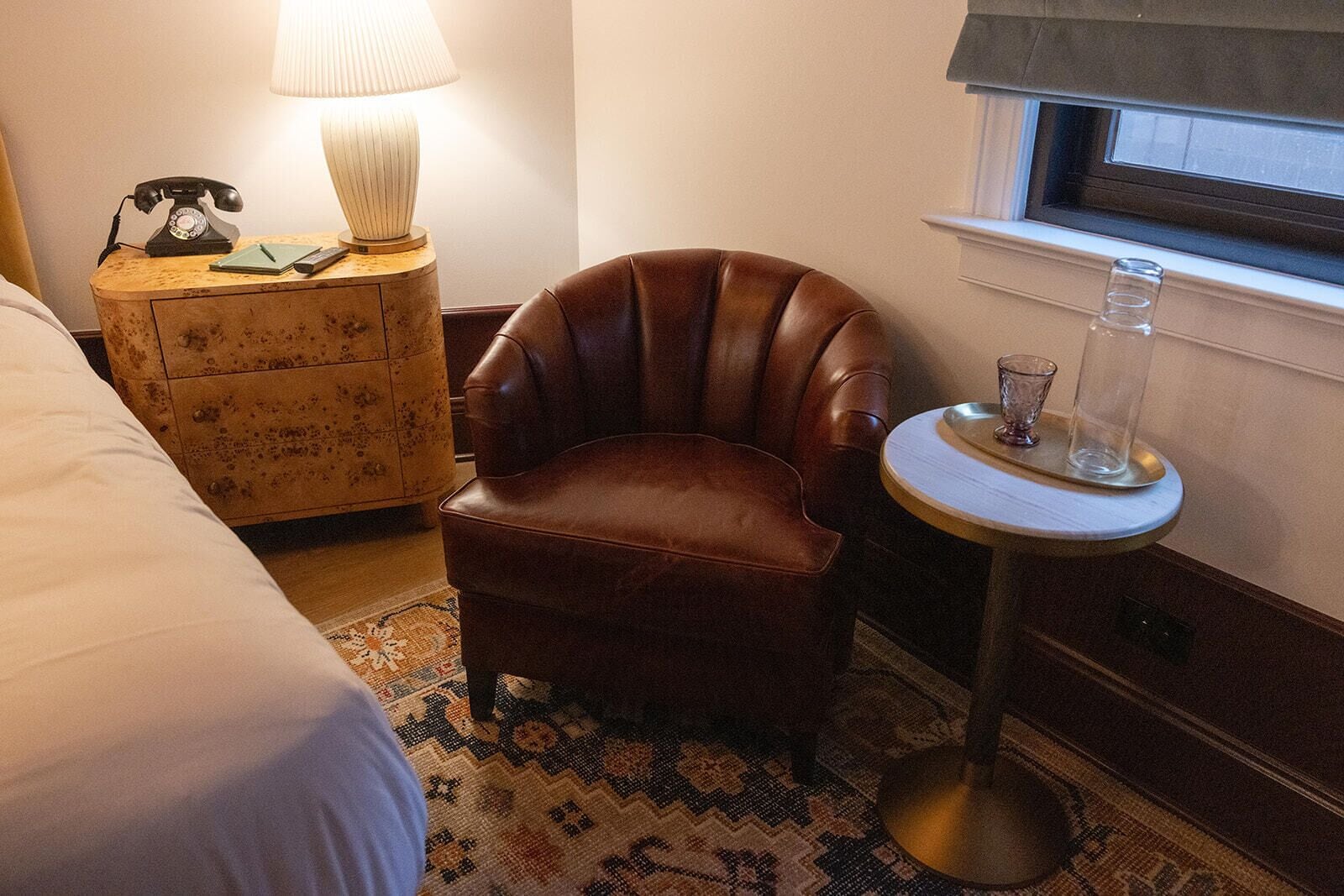 A hotel room with a brown leather armchair, a bedside table with a lamp, a telephone, and a window with a view.