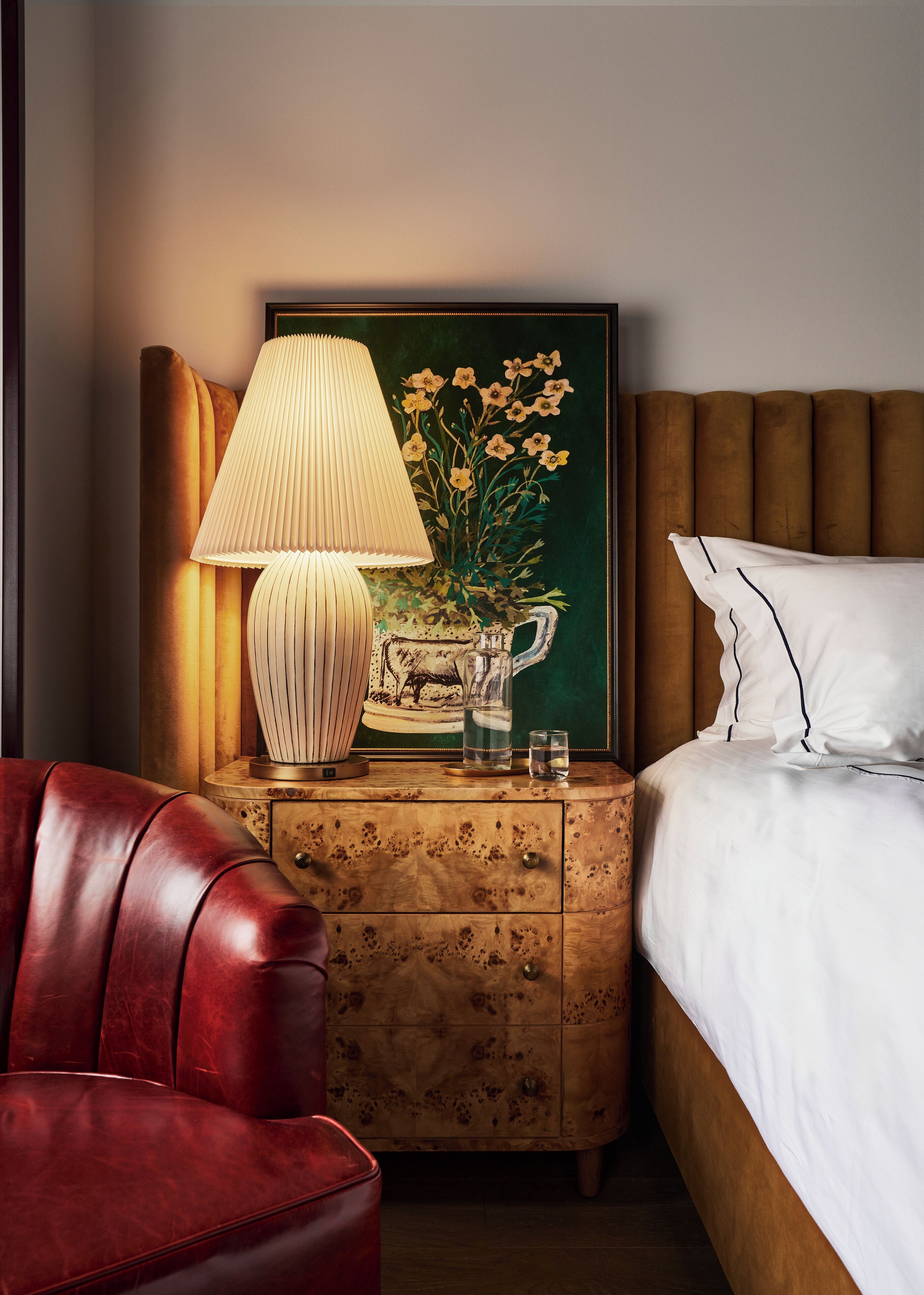 Un lit bien fait, avec du linge de lit blanc et un oreiller à rayure noire, un fauteuil en cuir rouge, une table de chevet en bois avec une lampe et un tableau de fleurs, et une tête de lit en bois.
