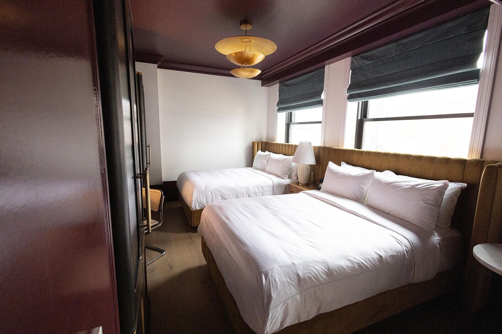 A hotel room with two beds, a wooden headboard, a window with blinds, a lamp, and a chair.
