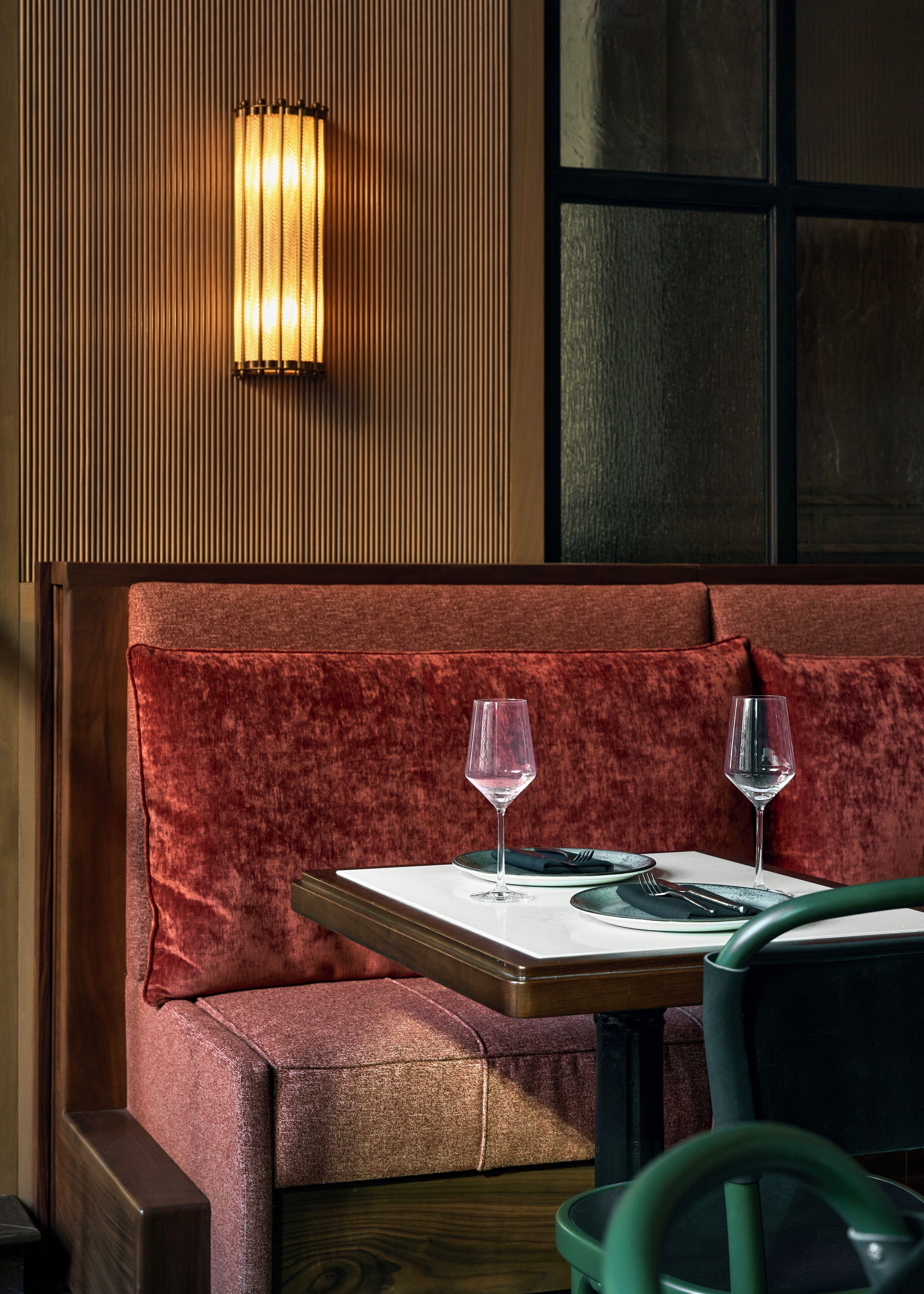 A restaurant seating area with a booth, two wine glasses, a table set with cutlery, and a wall-mounted light fixture.