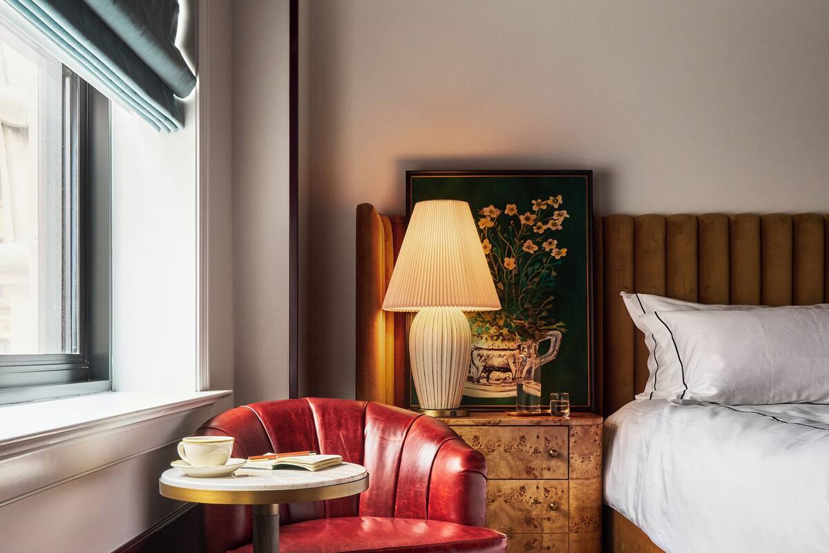 Habitación de hotel con una butaca de cuero rojo, una mesa redonda con una taza y un libro, una lámpara de noche, un cuadro enmarco y una cama con sábanas blancas.