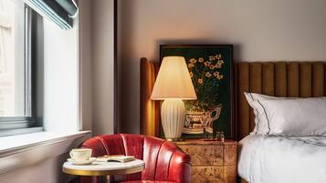 Hotelkamer met een rode lederen fauteuil, een rond tafeltje met een kop en boek, een bedlamp, een lijst met afbeelding en een bed met witte lakens.