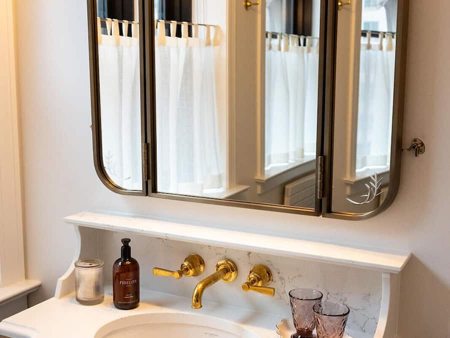 Un baño con un espejo grande, un lavabo blanco, grifos dorados y una botella de jabón Fidelity.