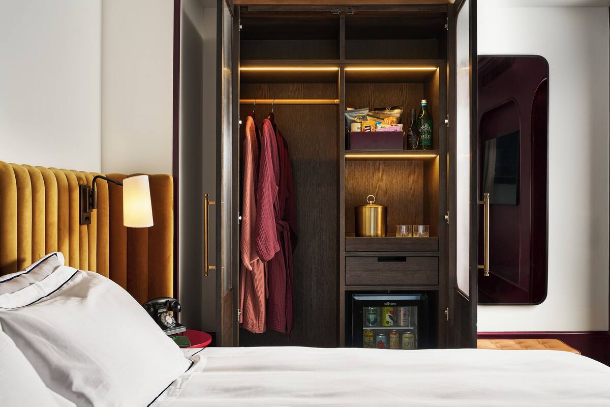 Una habitación de hotel con cama, mesita de noche con lámpara, armario con espejo y minibar.