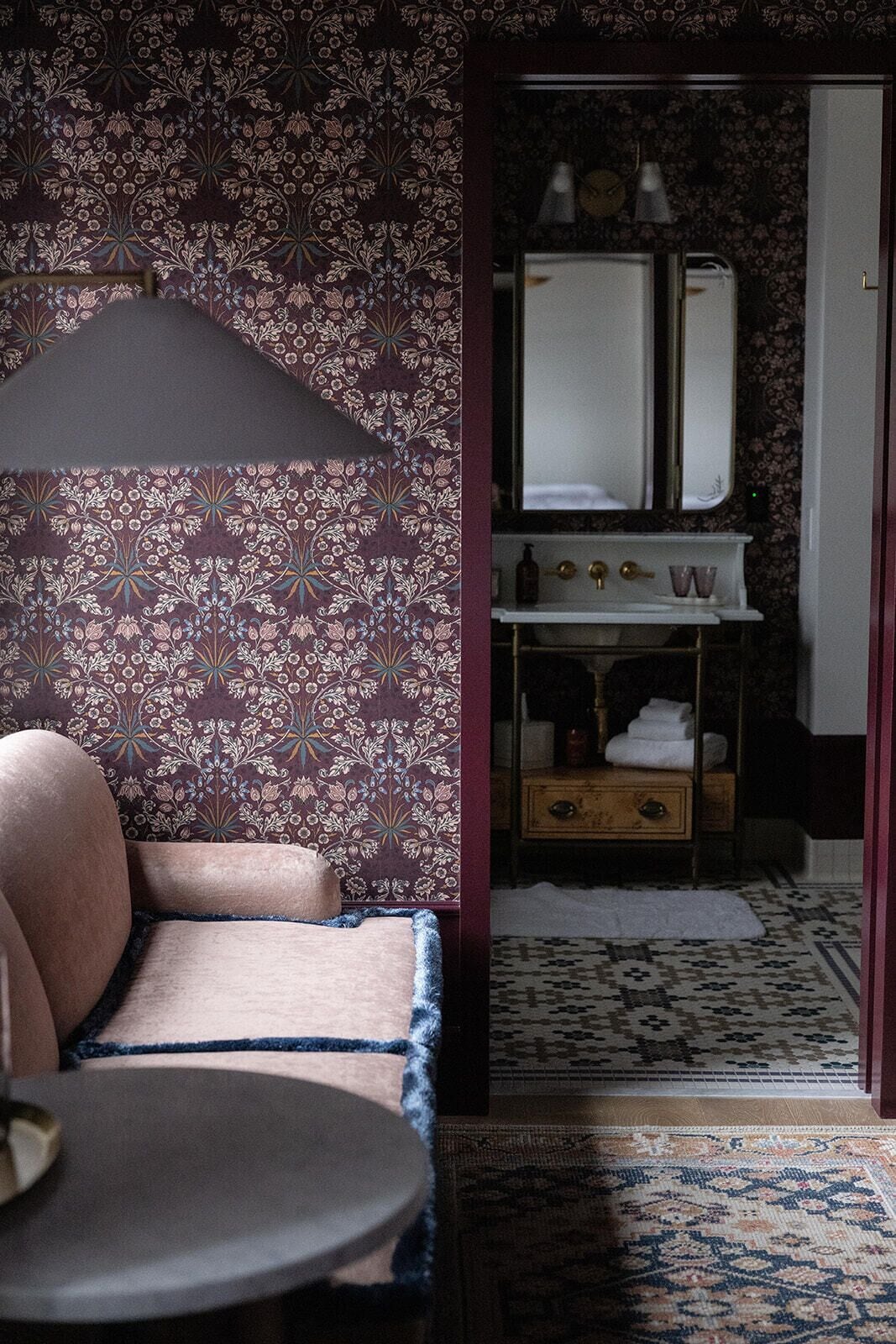 Een kamer met behang met patroon, een bank, een kleine tafel, een spiegel, een wastafel en een tapijt met patroon.