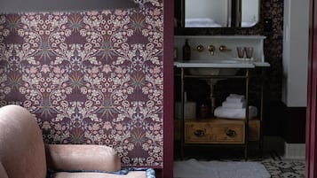 Een kamer met behang met patroon, een bank, een kleine tafel, een spiegel, een wastafel en een tapijt met patroon.
