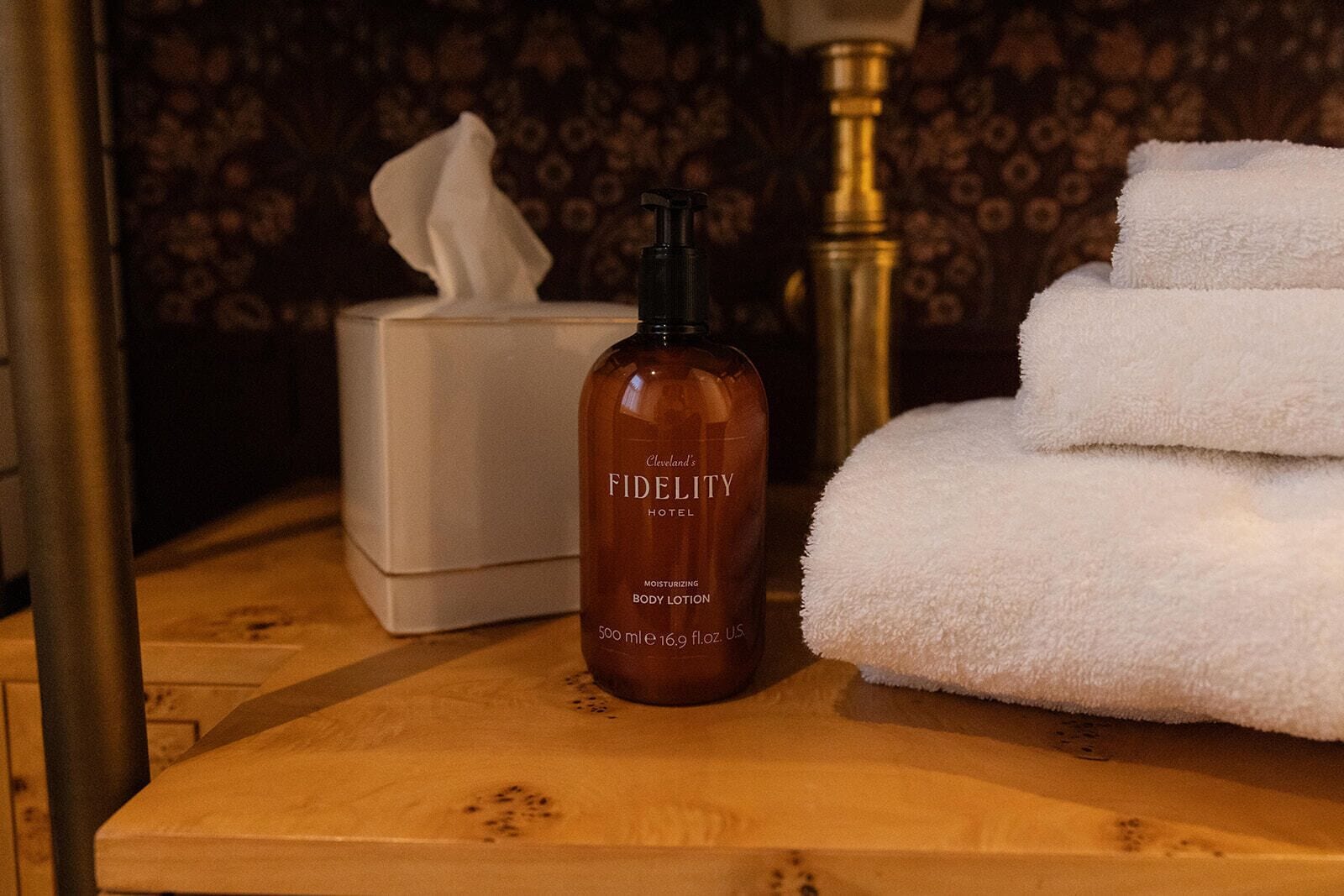 A bottle of Fidelity Hotel body lotion, a tissue box, and folded towels on a wooden surface.