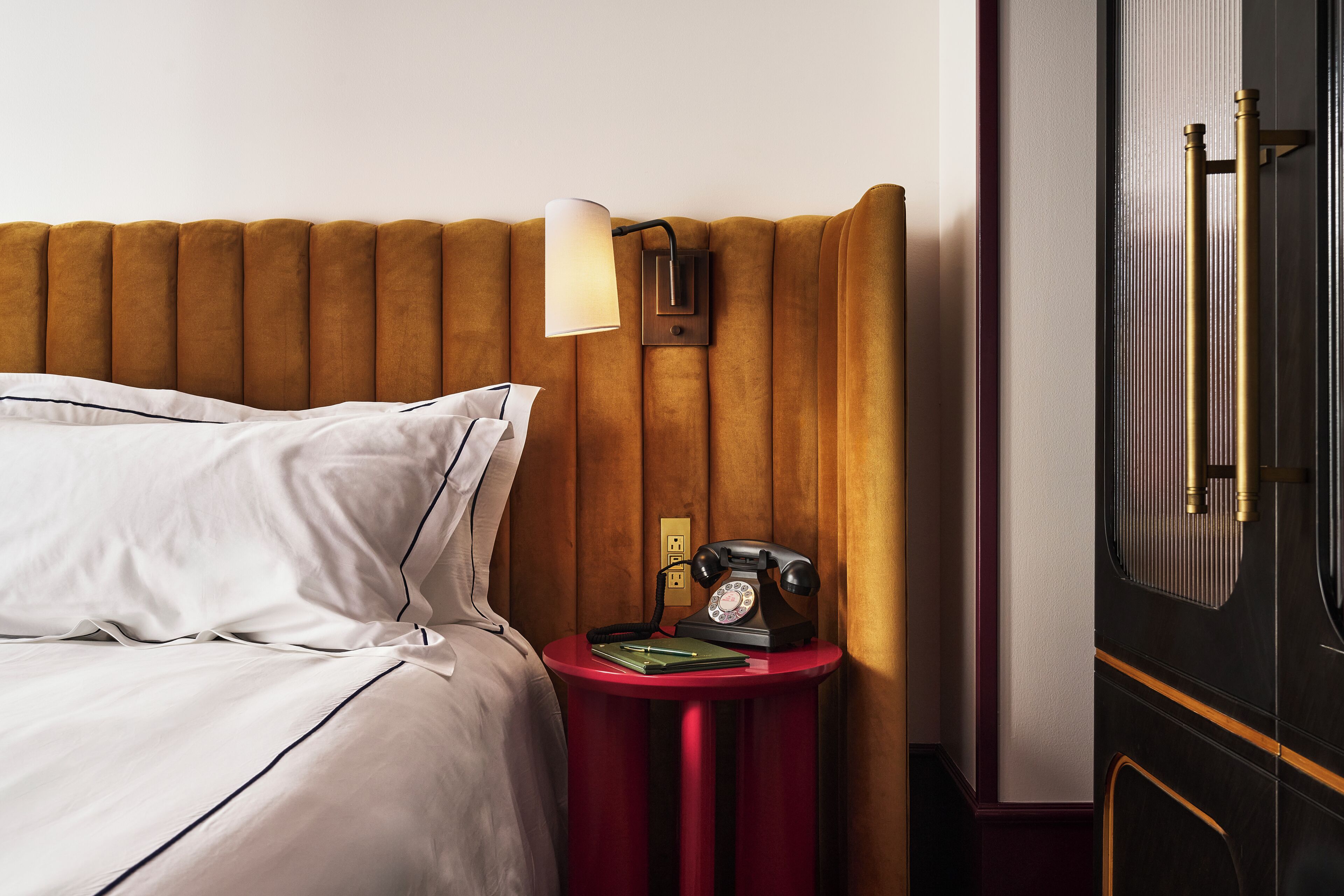 A neatly made bed with white linens, a red bedside table with a lamp, a telephone, and a green folder.