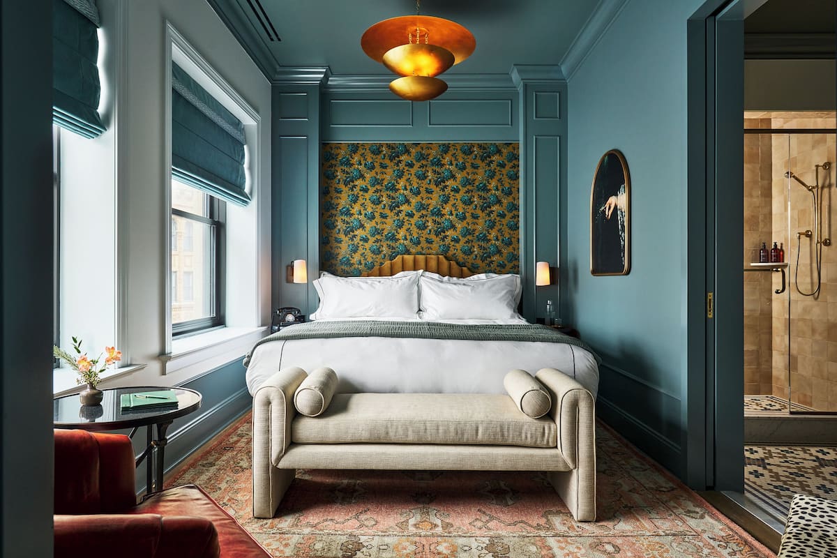 Un dormitorio con una cama grande, un banco, una pequeña mesa con flores y vistas al baño.