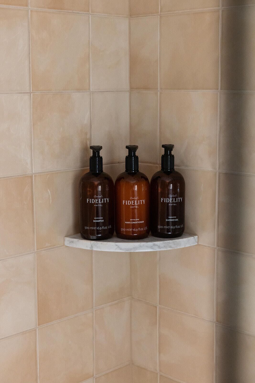 Three bottles of Fidelity Hotel shampoo, conditioner, and shower gel on a white shelf against a tiled wall.