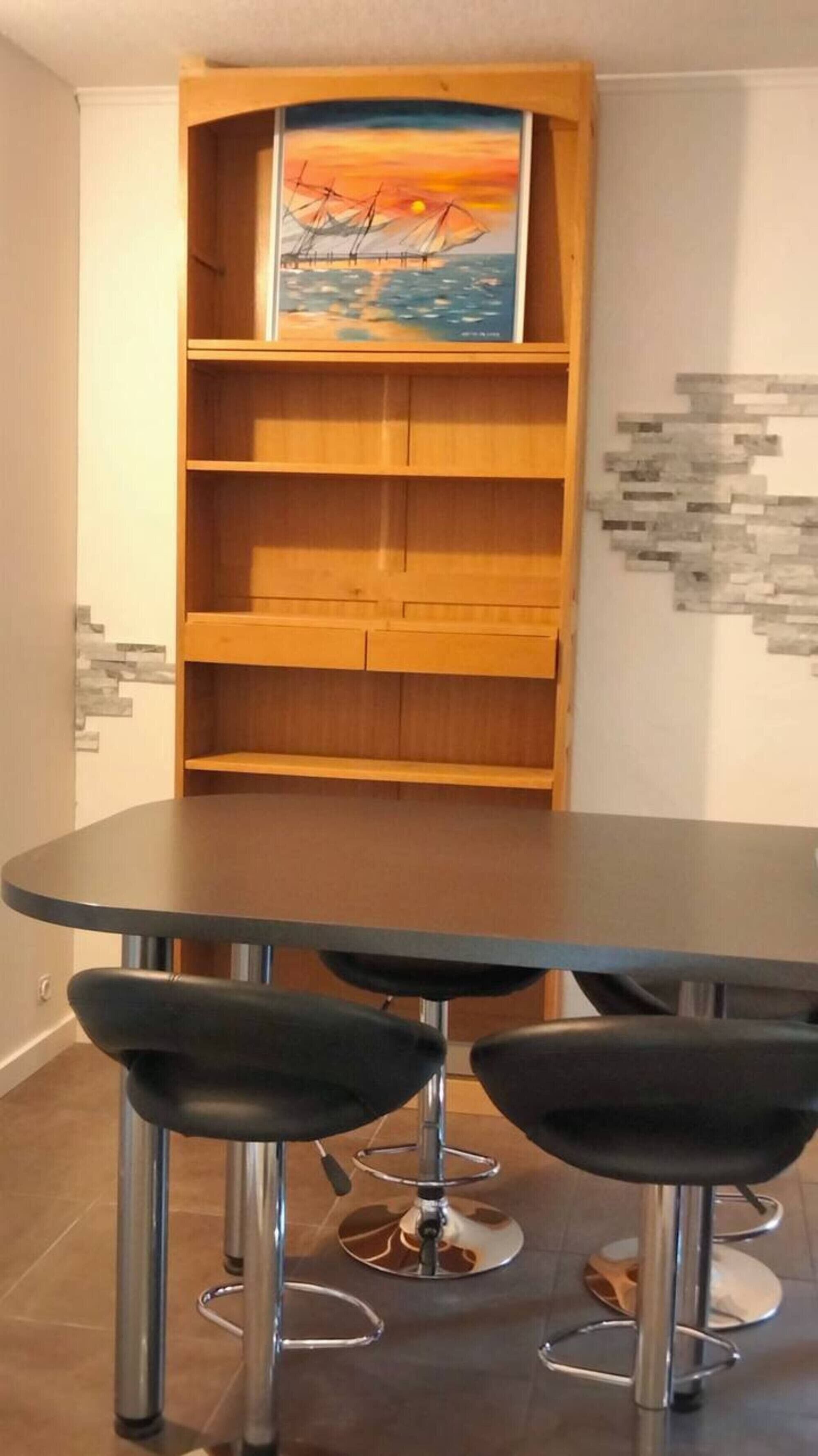 Un coin repas avec une table ronde, trois chaises noires et une bibliothèque en bois ornée d’un tableau.
