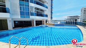 A swimming pool with a blue checkered pattern, surrounded by a building with balconies and a view of the sea. - PARCEROS GROUP - 2 BEDROOM VACATION APARTMENT, 6TH FLOOR, CARTAGENA, SPAIN (Cartagena de Indias)