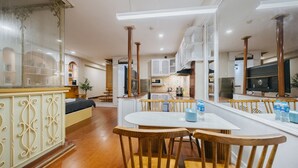 A spacious dining area with a white table and wooden chairs, a kitchen in the background, and a large mirror reflecting the room.