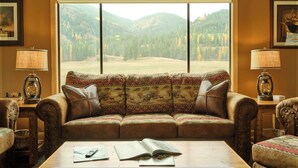 A cozy living room with a leather sofa, a coffee table, and a large window offering a view of a forested mountain. - Black Bear Lodge - Alpine Falls Ranch (Superior)
