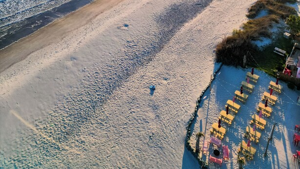 Vista aérea de una playa con un muelle que se adentra en el agua, cielos despejados y mar tranquilo.
