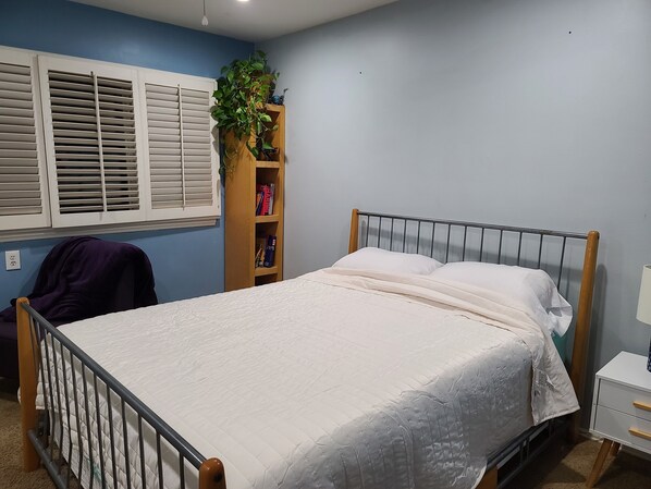Un dormitorio con cama, mesita de noche de madera, estantería y ventana con persianas.