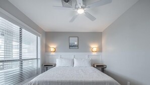 Une chambre à coucher avec un grand lit, deux tables de chevet, un ventilateur de plafond et une fenêtre avec des stores.