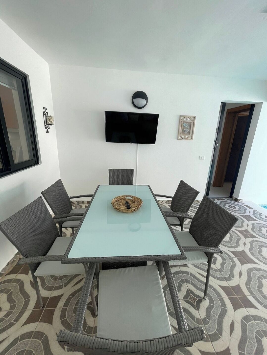 A modern room with a glass-top table, wicker chairs, a flat-screen TV mounted on the wall, and patterned floor tiles.