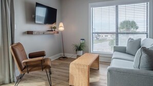 Una sala de estar moderna con un sofá gris, un sillón de cuero marrón, un banco de madera, un televisor de pantalla plana en la pared y un ventanal con persianas.