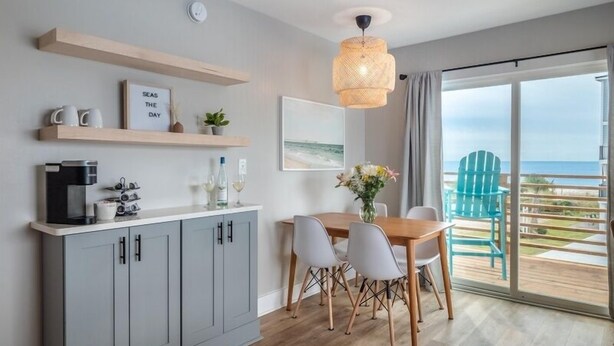 Una cocina moderna con armarios grises, una máquina de café y un comedor con mesa y sillas de madera. Se ve el océano a través de ventanas amplias.