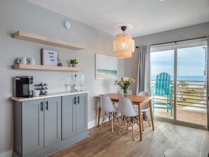 Uma cozinha moderna com armários cinza, cafeteira e área de refeições com mesa e cadeiras de madeira. Há vista para o oceano através de grandes janelas.