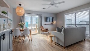 Una sala de estar moderna con un sofá gris, piso de madera, un comedor con sillas blancas, un ventilador de techo y un televisor montado en la pared.