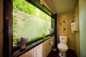 Un baño con ventana, paneles de madera, inodoro, cubo de basura y una planta pequeña en el alféizar.