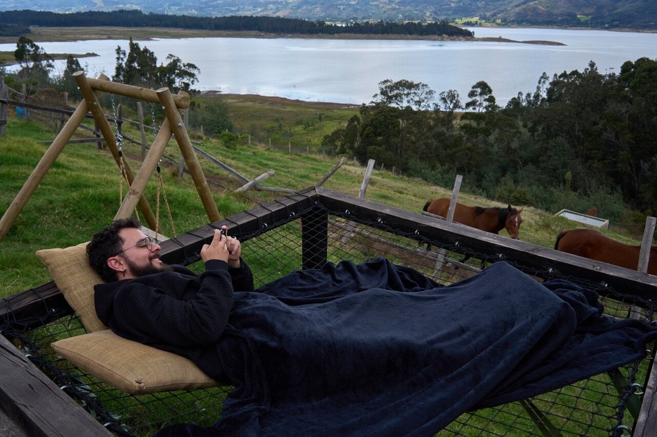 Una persona recostada en una silla hamaca, con vista a un lago y caballos al fondo.