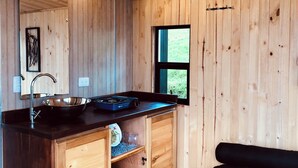 Een kleine keuken met een spoelbak, fornuis en houten kastjes. Een raam zorgt voor daglicht en uitzicht op groen.