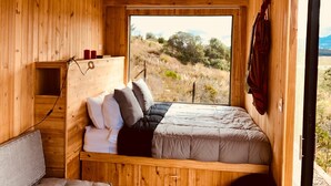 Een houten slaapkamer met een bed, kussens, een plank met kaarsen en een raam met uitzicht op een landschap.