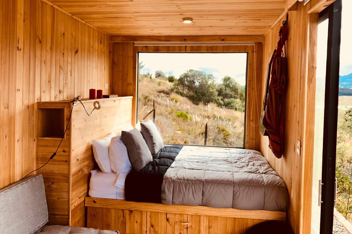 A wooden cabin bedroom with a bed, pillows, a shelf with candles, and a window overlooking a landscape.
