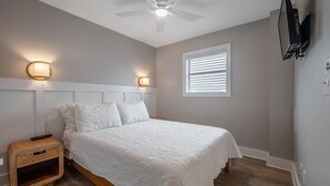 Quarto com cama, abajures, ventilador de teto, janela com persianas e televisão fixada na parede.