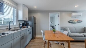 Una cocina moderna con horno empotrado, un lavamanos y una ventana con persianas. Hay un comedor con una mesa y sillas de madera, y un sofá gris en la sala de estar.