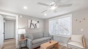 A modern living room with a grey sofa, a wooden coffee table, a white armchair, a ceiling fan, and a window with blinds. - Beach House OKI - Queen Suite in hotel on Oak Island, North Carolina (Oak Island)