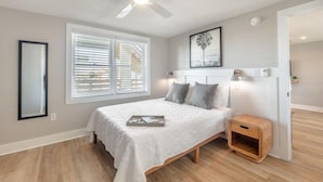 A bedroom with a bed, a ceiling fan, a window with blinds, a framed picture on the wall, and a small wooden nightstand. - Beach House OKI - Queen Suite in hotel on Oak Island, North Carolina (Oak Island)