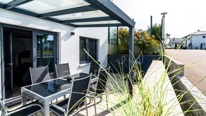 An outdoor patio with a glass table and chairs, a sliding glass door, and a view of the street. - ALP DELUXE - Family-friendly and quiet vacation apartment with sauna & terrace (Weißenhorn)