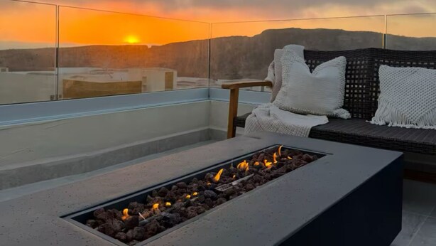 Une terrasse sur le toit dotée d’un foyer, d’un espace salon avec des coussins et offrant une vue sur le coucher de soleil au-dessus de la ville.
